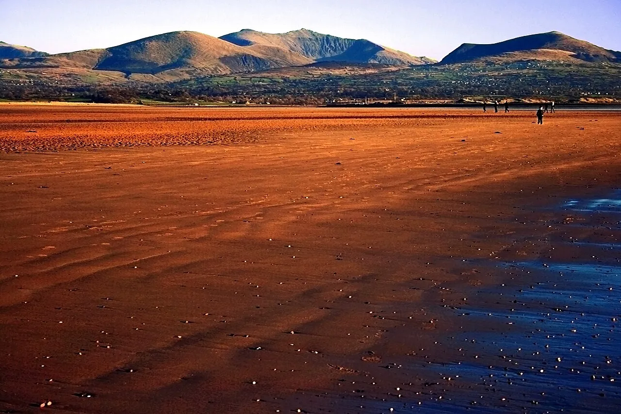 Ynys Llanddwyn beach at low tide, wide amber sand stretching towards Snowdonia's mountains across the water.