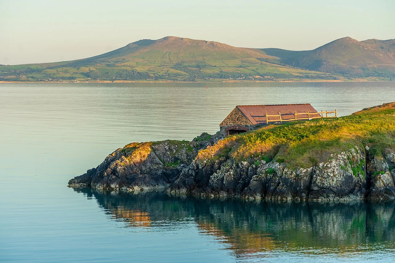 A stone boathouse on the rocky shore of Llanddwyn, with the mountains of the Llŷn Peninsula across calm water at golden hour.