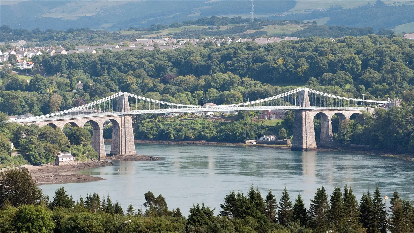The full span of the Menai Suspension Bridge seen from the Marquess of Anglesey Column, the road deck crossing the strait between wooded banks.