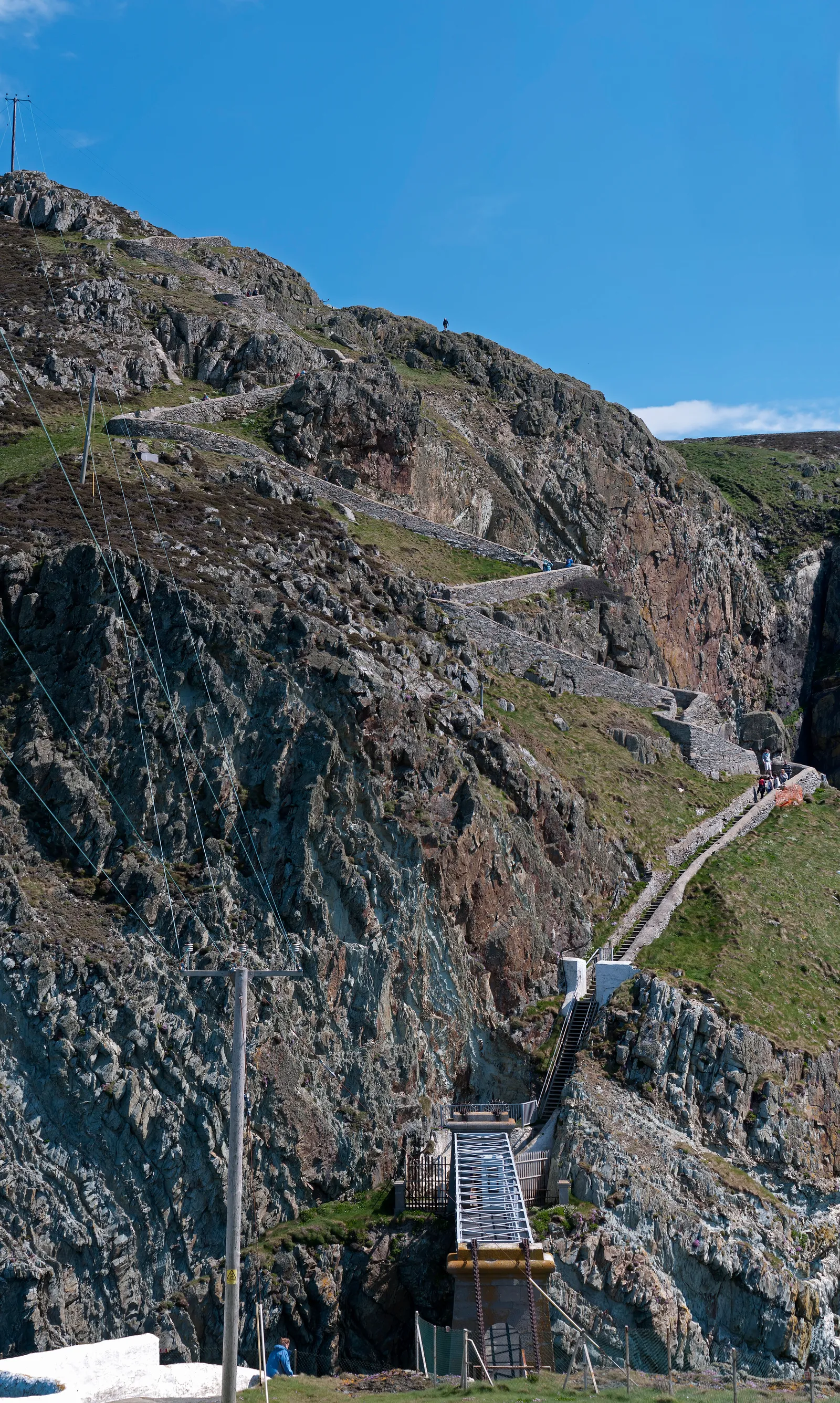 The steep zig-zag path and white footbridge dropping down the cliff towards South Stack Lighthouse island, walkers small on the steps.